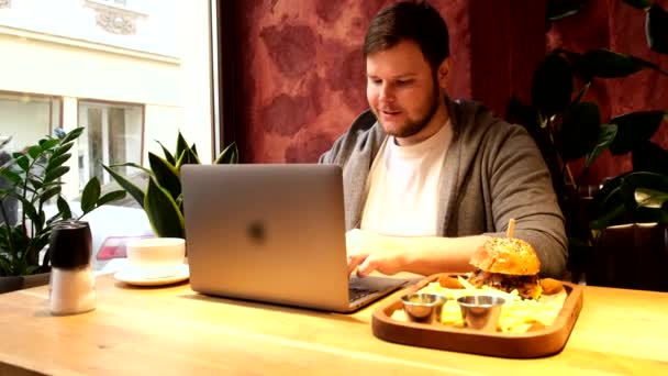 Homme travaillant sur ordinateur portable dans un café manger un hamburger — Video