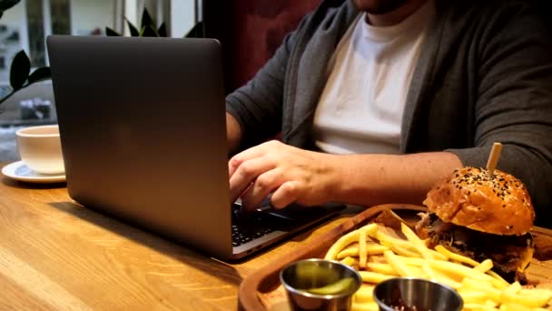 カフェでノートパソコンでバーガーを食べる男 — ストック動画