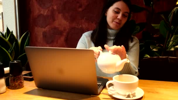 在咖啡店的笔记本电脑上工作的女自由职业者用杯子倒茶 — 图库视频影像