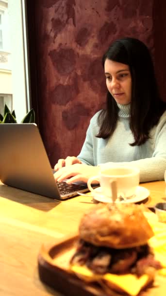 Žena pracující na dálku na notebooku v kavárně jíst burger pití čaje — Stock video