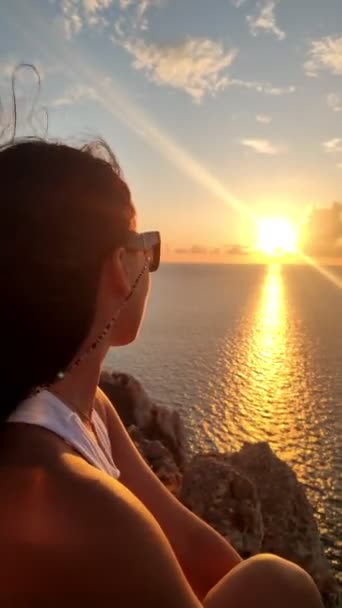 Mulher sentada no penhasco desfrutando do pôr do sol acima do mar — Vídeo de Stock