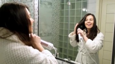 Mutlu kadın banyoda aynanın önünde saçlarını kurutup şarkı söylüyor.