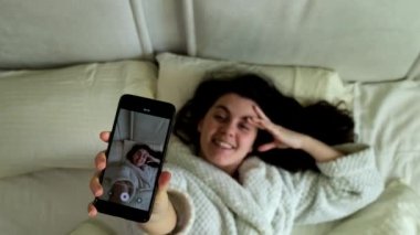 Mutlu gülümseyen blogcu yatakta yatarken telefonda video çekiyor.