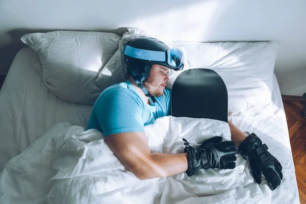 Kar kayağı google 'ı ve kaskla yatakta kar dağlarını düşleyen bir adam.