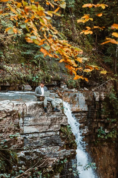 Jovem homem forte trabalhando no laptop sentado à beira da cachoeira — Fotografia de Stock