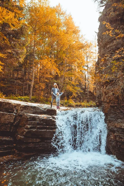 Jonge vrouw met peuter meisje kijken naar waterval in de herfst bos — Stockfoto