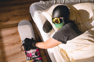 Kayak ekipmanlı bir adam, dağları düşleyen bir snowboard 'la yatakta uyuyor.