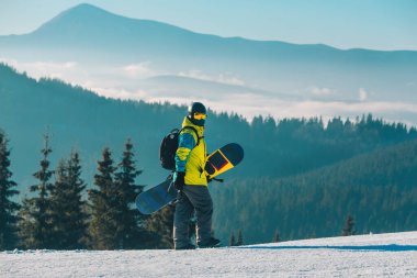 Arka planda snowboard dağlarıyla tepenin yanından geçen bir adam.