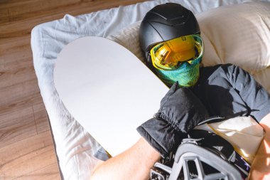 Kayak ekipmanlı bir adam, dağları düşleyen bir snowboard 'la yatakta uyuyor.