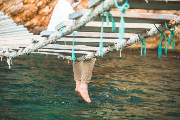Gewas foto man zittend op hangbrug zet benen in zeewater — Stockfoto