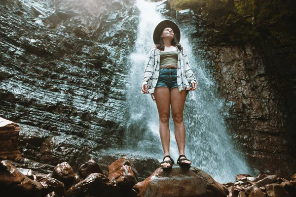 Kahverengi şapkalı kadın şelale yürüyüşünün tadını çıkarıyor.