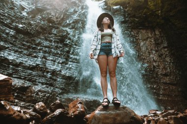 Kahverengi şapkalı kadın şelale yürüyüşünün tadını çıkarıyor.