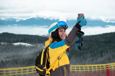 Kadın kayakçı arka plandaki telefon dağlarında selfie çekiyor.