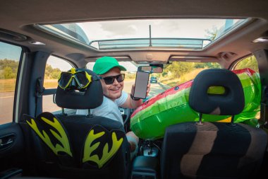 Mutlu sürücü arabası dolusu tatil malzemesi. Telefonu beyaz ekran fotokopi alanı ile tutuyor.