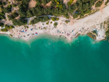 Mavi gök mavisi suda yüzen, kumlu sahilde güneşlenen insanların havadan görüntüsü.