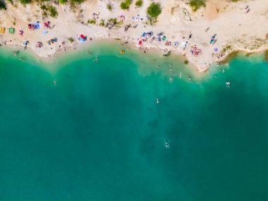 Mavi gök mavisi suda yüzen, kumlu sahilde güneşlenen insanların havadan görüntüsü.