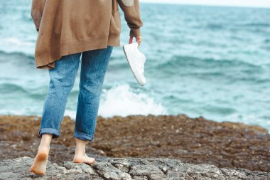 Deniz kenarında çıplak ayakla yürüyen bir kadın el ele tutuşmuş beyaz ayakkabılar giyiyor. Yaz tatili. Doğa birliği