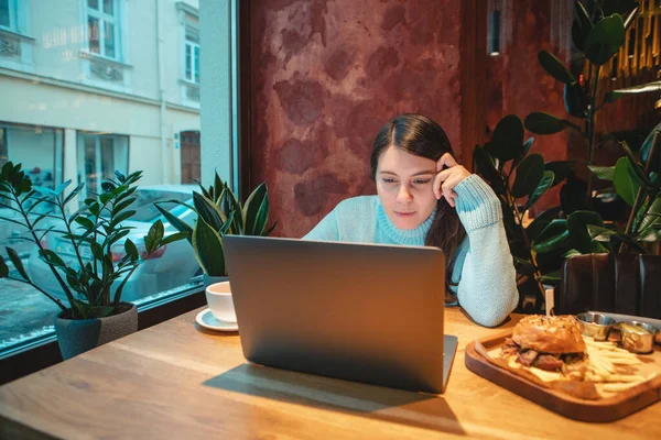 在咖啡店的笔记本电脑上工作的女自由职业者喝茶 吃汉堡包 复印空间 — 图库照片