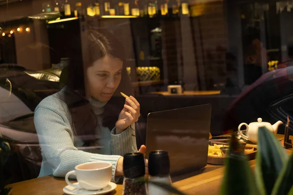 在餐馆里用笔记本电脑工作的微笑的女人 一边喝茶一边吃汉堡包 透过玻璃杯观看 — 图库照片