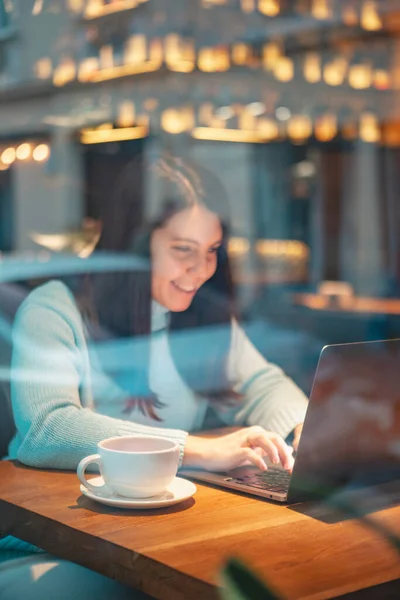 レストランでノートパソコンで働く笑顔の女性がガラス越しにバーガービューを食べる — ストック写真