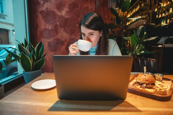 在咖啡店的笔记本电脑上工作的女自由职业者喝茶 吃汉堡包 复印空间 — 图库照片
