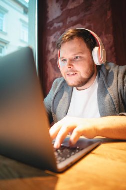 Adam kulaklık takmış, laptopla kafede çalışıyor, hamburger yiyor ve çay fotokopisi çekiyor.