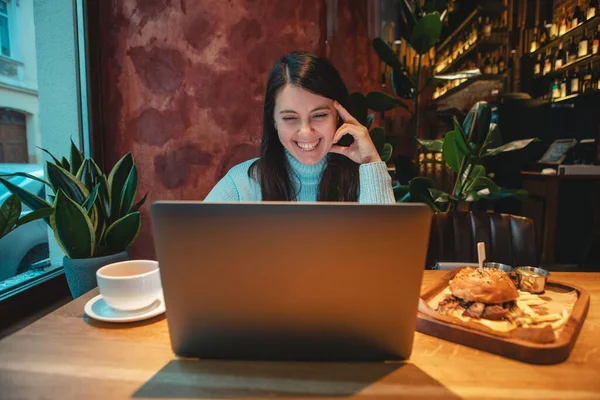 Woman freelancer working on laptop in cafe drinking tea eating burger — Stock Photo, Image