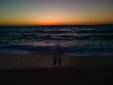 Deniz kıyısında durup gün batımında güçlü dalgalara bakan çiftlerin silueti.