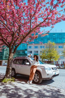 Gülümseyen güzel bir kadın arabanın yanında duruyor. Arka planda çiçek açan sakura ağacı. Bahar geliyor.
