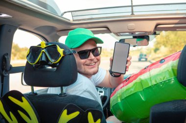 Mutlu sürücü arabası dolusu tatil malzemesi. Telefonu beyaz ekran fotokopi alanı ile tutuyor.