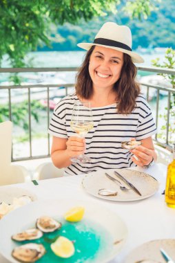 Fötr şapkalı, açık hava restoranında oturmuş istiridye yiyip şarap içen gülümseyen güzel kadın. yaz zamanı