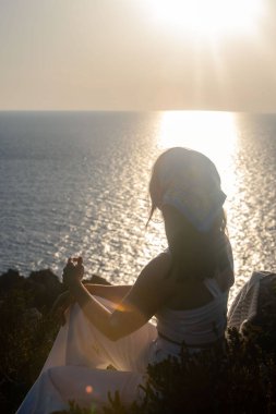 Uçurumda oturan kadın yaz mevsiminde gün batımını seyrediyor.