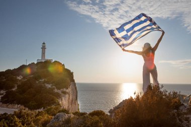 Yunan bayrağı taşıyan kadın arka planda Lefkada adasının deniz fenerinde gün batımına bakıyor.
