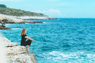 Genç, sıska bir kadın uçurumun kenarında oturmuş denize bakıyor. Boşluğu kopyala