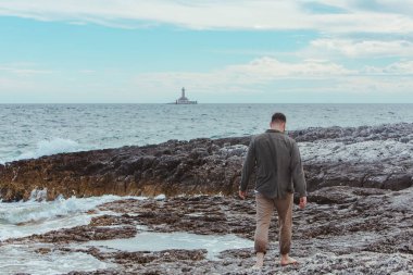 Genç bir adam kayalık deniz plajında çıplak ayakla yaz tatilinde yürüyor. Arka planda deniz feneri