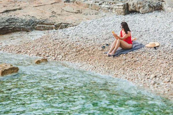 Mladá Hezká Žena Červených Plavkách Sedí Skalnaté Pláži Telefonem Moře — Stock fotografie