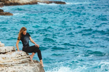 Genç, sıska bir kadın uçurumun kenarında oturmuş denize bakıyor. Boşluğu kopyala