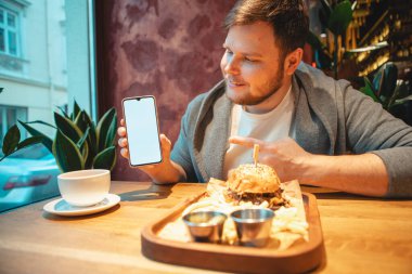 Kafede elinde telefonla hamburger yiyip çay içen bir adam.