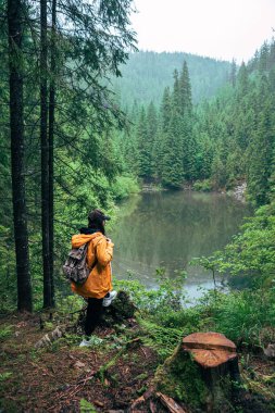 Sırt çantalı, sarı yağmurluklu bir kadın dağ gölü fotokopi alanına bakıyor.