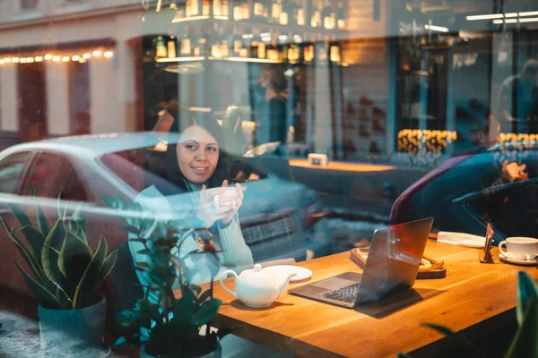 在餐馆里用笔记本电脑工作的微笑的女人 一边喝茶一边吃汉堡包 透过玻璃杯观看 — 图库照片