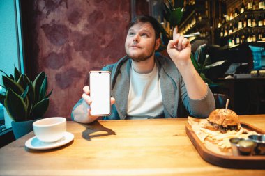 Kafede elinde telefonla hamburger yiyip çay içen bir adam.