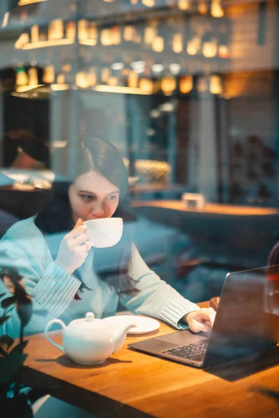 smiling woman working on laptop in restaurant drinking tea eating burger view through the glass