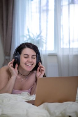 woman laying down in bed working on laptop listening music in headset bright white room