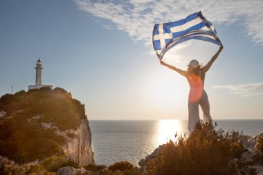 Yunan bayrağı taşıyan kadın arka planda Lefkada adasının deniz fenerinde gün batımına bakıyor.