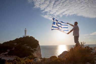 Yunan bayraklı bir adam arka planda Lefkada adasının deniz fenerine bakıyor.