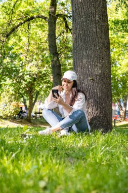 Kadın, şehir parkındaki ağacın altında oturmuş telefon fotokopi odasında internette sörf yapıyor.