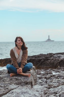 arka planda ıslak kot deniz feneri kayalık deniz plajı nda oturan kadın. rüzgarlı hava. yaz tatili. kaygısız kavram
