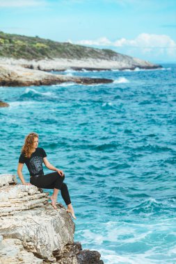 Genç, sıska bir kadın uçurumun kenarında oturmuş denize bakıyor. Boşluğu kopyala
