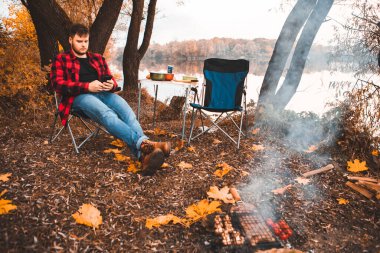 man sitting near bonfire surfing internet on the phone autumn camping concept