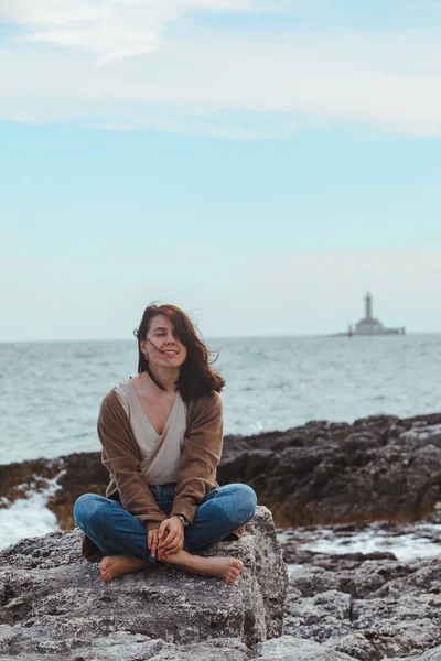 woman sitting by rocky sea beach in wet jeans lighthouse on background. windy weather. summer vacation. carefree concept
